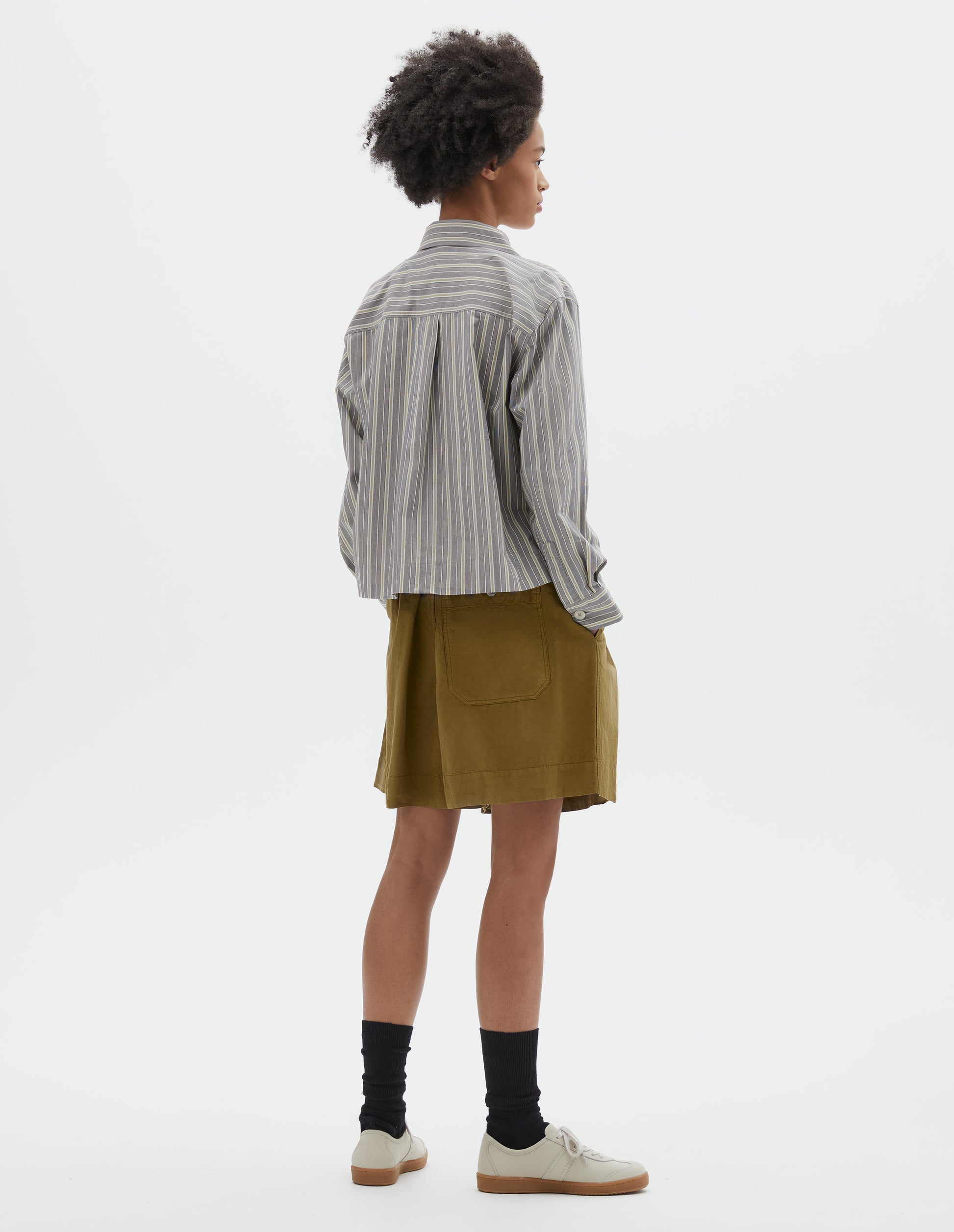 Full body image, back view of model wearing grey, yellow and white striped cotton long sleeve shirt. With stepped vent split hem, corozo brace button cuffs and box pleat on back yoke. Styled with pleated green shorts, black socks and beige suede trainers.