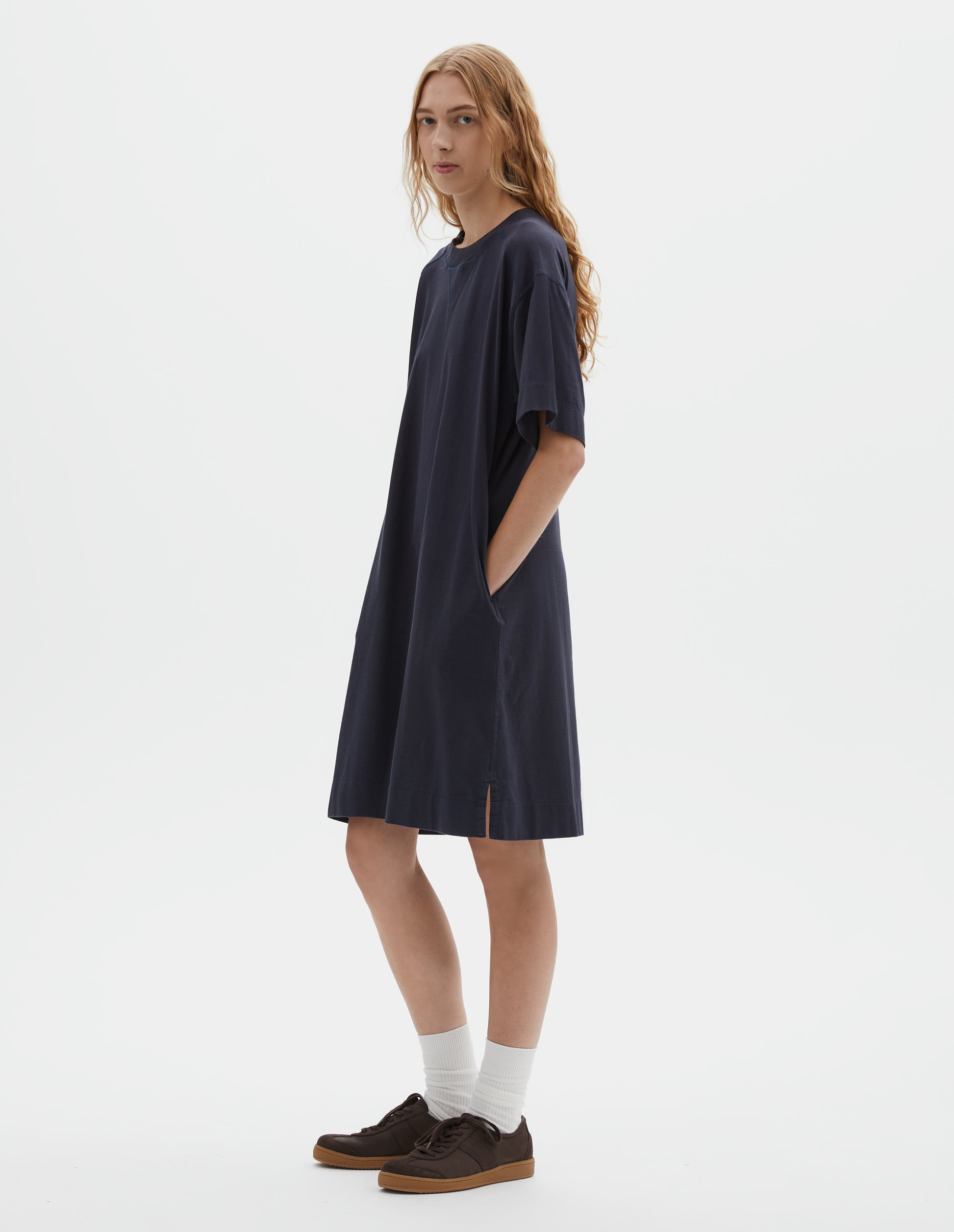 Full body image, front view of model wearing dark navy blue short sleeve cotton t-shirt dress, with concealed side pockets and seam detailing, side split hems. Styled with white socks and brown suede trainers.