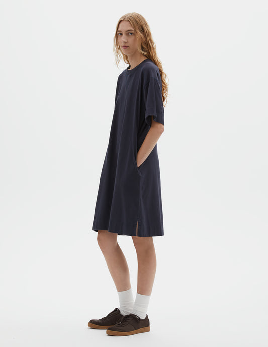 Full body image, front view of model wearing dark navy blue short sleeve cotton t-shirt dress, with concealed side pockets and seam detailing, side split hems. Styled with white socks and brown suede trainers.