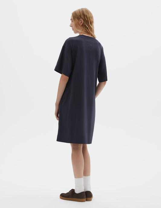 Full body image, back view of model wearing dark navy blue short sleeve cotton t-shirt dress, with seam detailing, and side split hems. Styled with white socks and brown suede trainers.