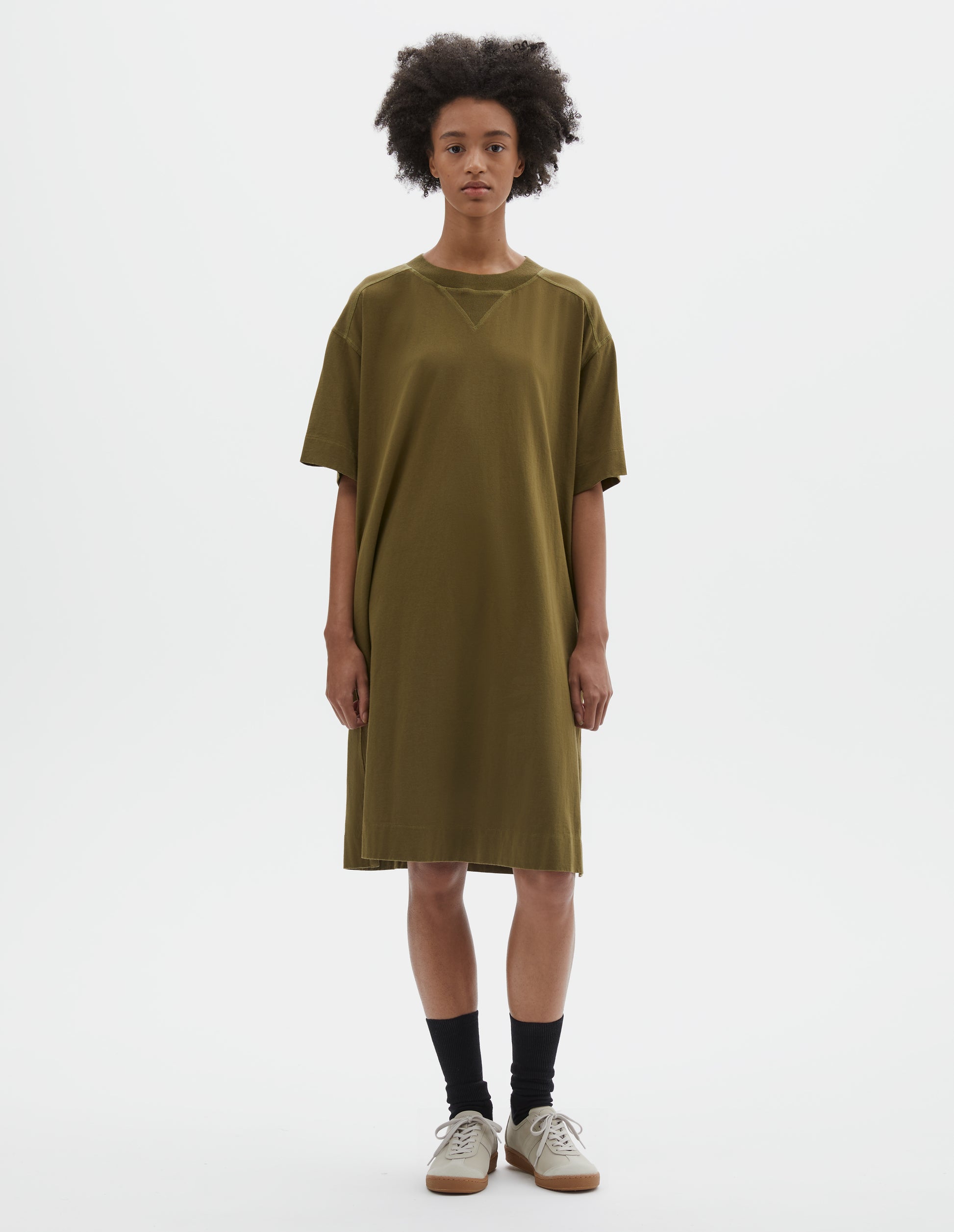 Full body image, front view of model wearing brownish green short sleeve cotton t-shirt dress. With concealed side pockets, side split hems and flatlock stitch seam detailing. Styled with black socks and beige suede trainers.