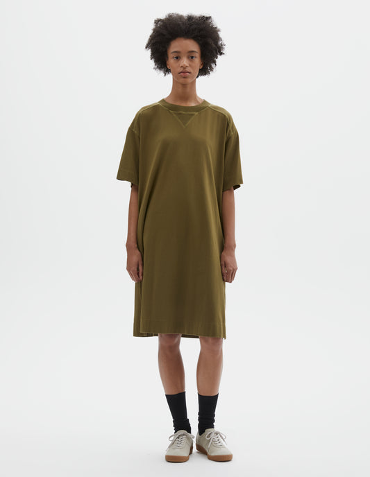 Full body image, front view of model wearing brownish green short sleeve cotton t-shirt dress. With concealed side pockets, side split hems and flatlock stitch seam detailing. Styled with black socks and beige suede trainers.