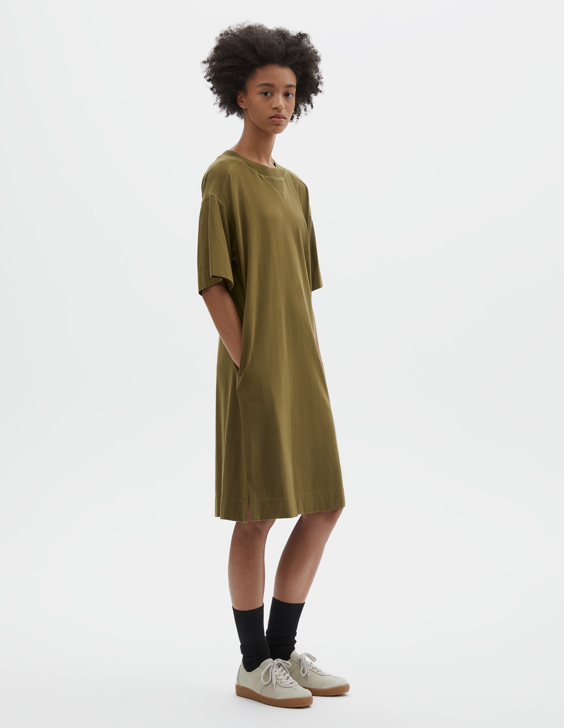 Full body image, side view of model wearing brownish green short sleeve cotton t-shirt dress. With concealed side pockets, side split hems and flatlock stitch seam detailing. Styled with black socks and beige suede trainers.