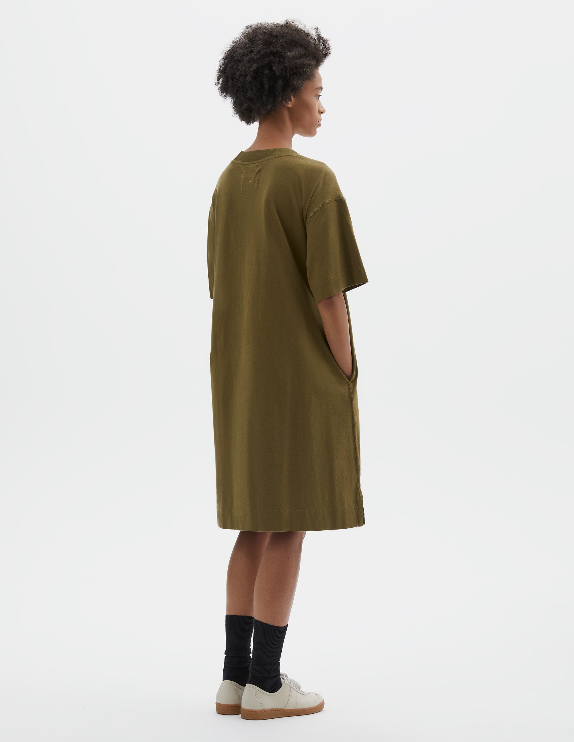 Full body image, back view of model wearing brownish green short sleeve cotton t-shirt dress. With concealed side pockets, side split hems and flatlock stitch seam detailing. Styled with black socks and beige suede trainers.