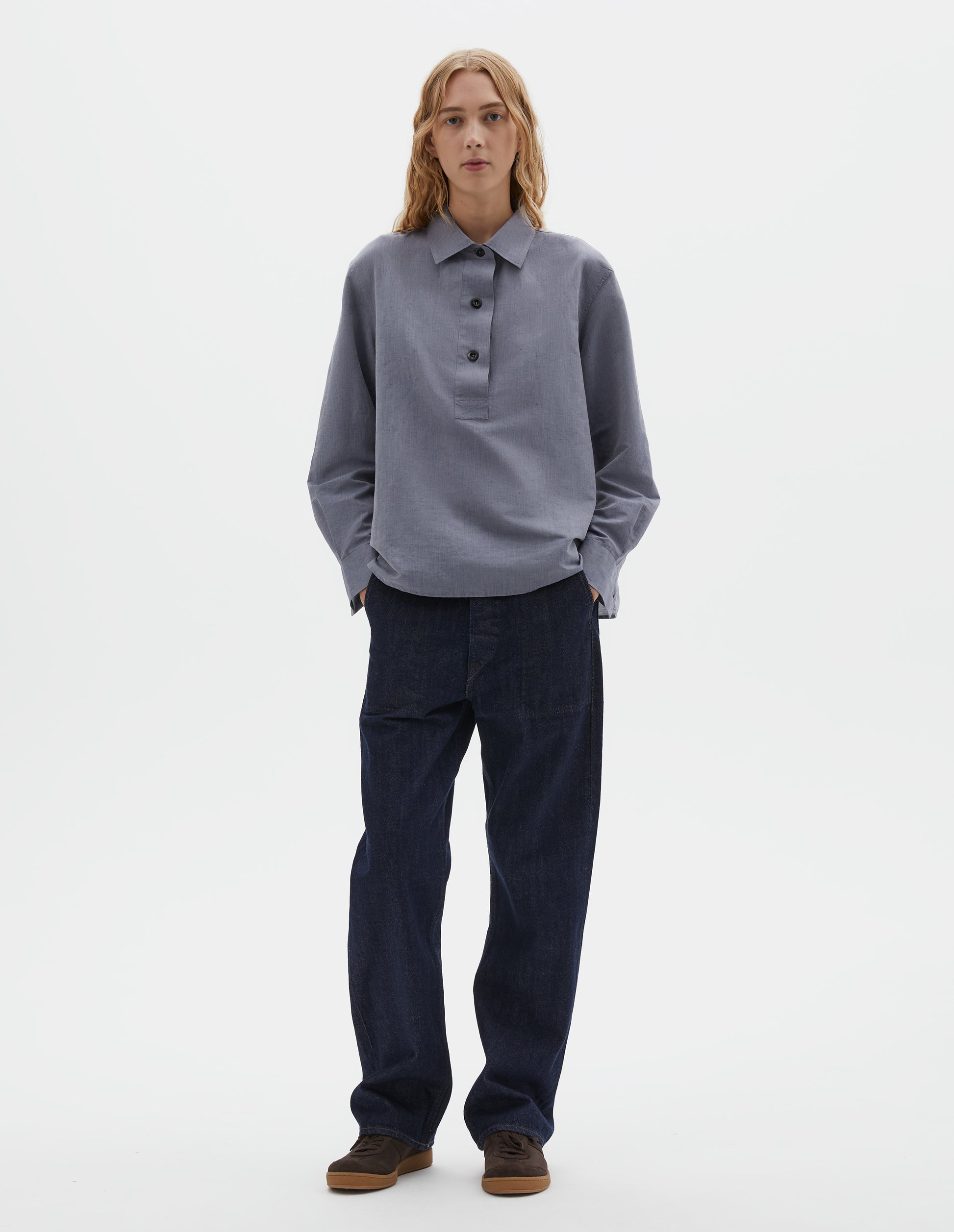 Full body image, front view of model wearing indigo blue denim twill jeans, with metal button fly, stitch-through waistband with belt loops, and stitch through pockets. Styled with blue chambray shirt and brown suede trainers.