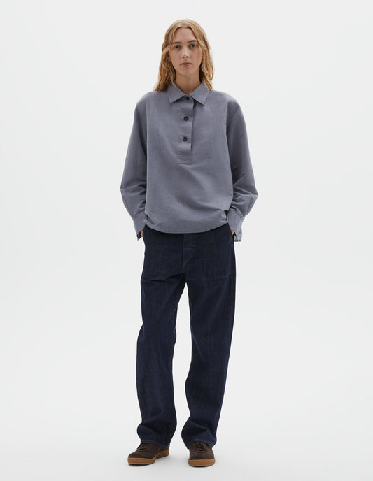 Full body image, front view of model wearing indigo blue denim twill jeans, with metal button fly, stitch-through waistband with belt loops, and stitch through pockets. Styled with blue chambray shirt and brown suede trainers.