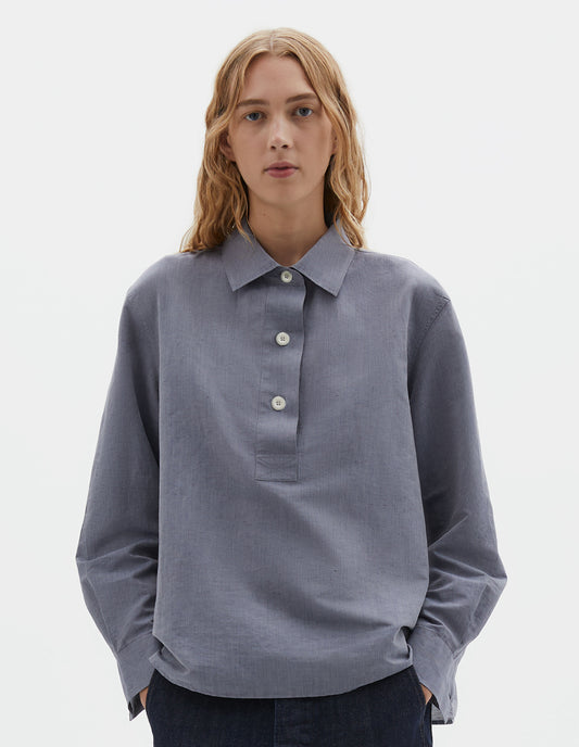 Cropped body image, front view of model wearing greyish blue long sleeve cotton linen shirt, with half raised placket and corozo brace buttons. Styled with indigo blue jeans.