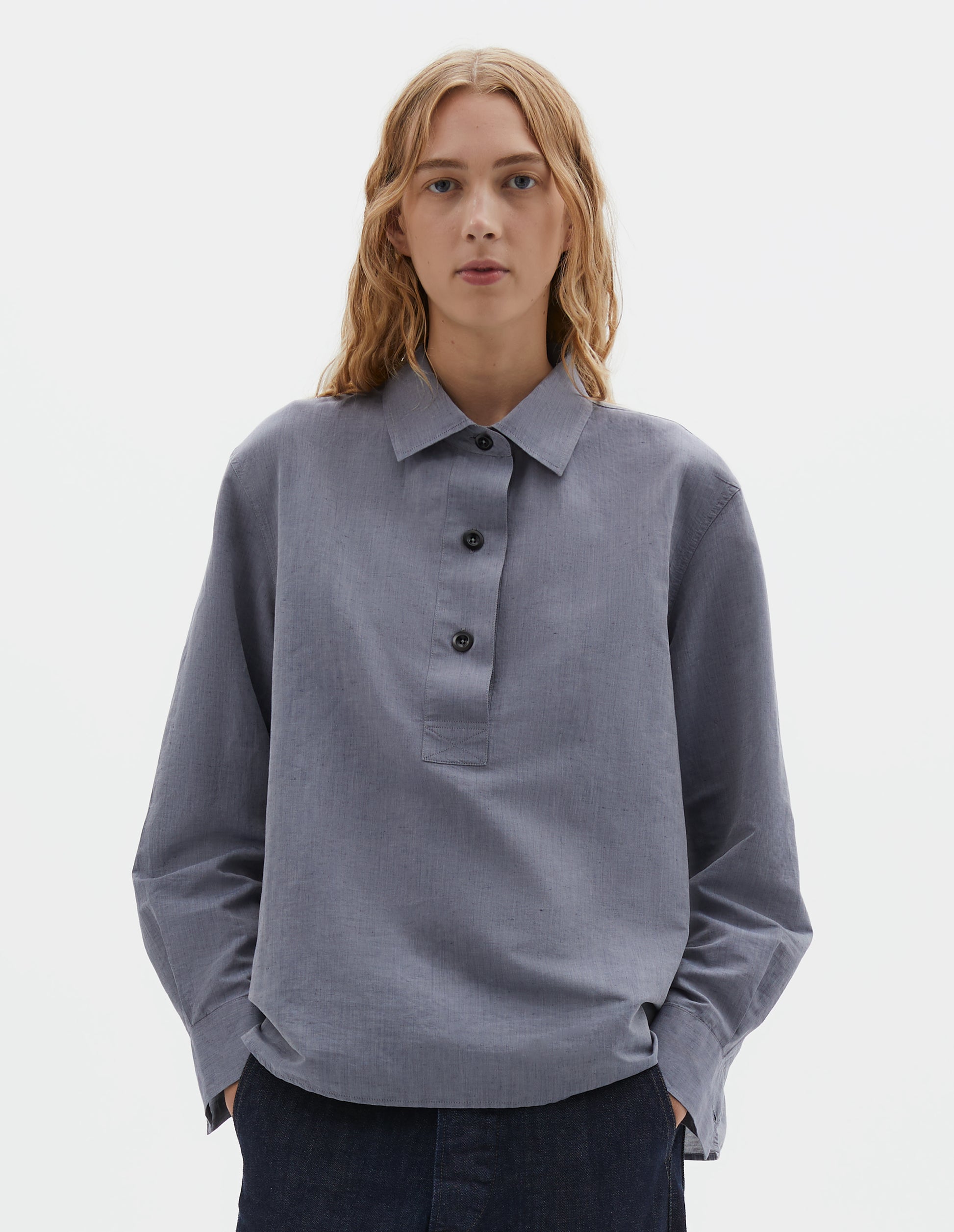Cropped body image, front view of model wearing greyish blue long sleeve cotton linen shirt, with half raised placket and corozo brace buttons. Styled with indigo blue jeans.