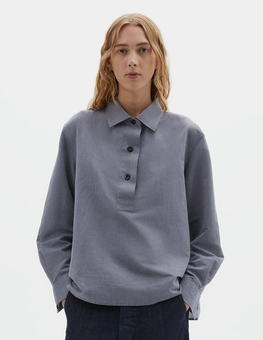 Cropped body image, front view of model wearing greyish blue long sleeve cotton linen shirt, with half raised placket and corozo brace buttons. Styled with indigo blue jeans.