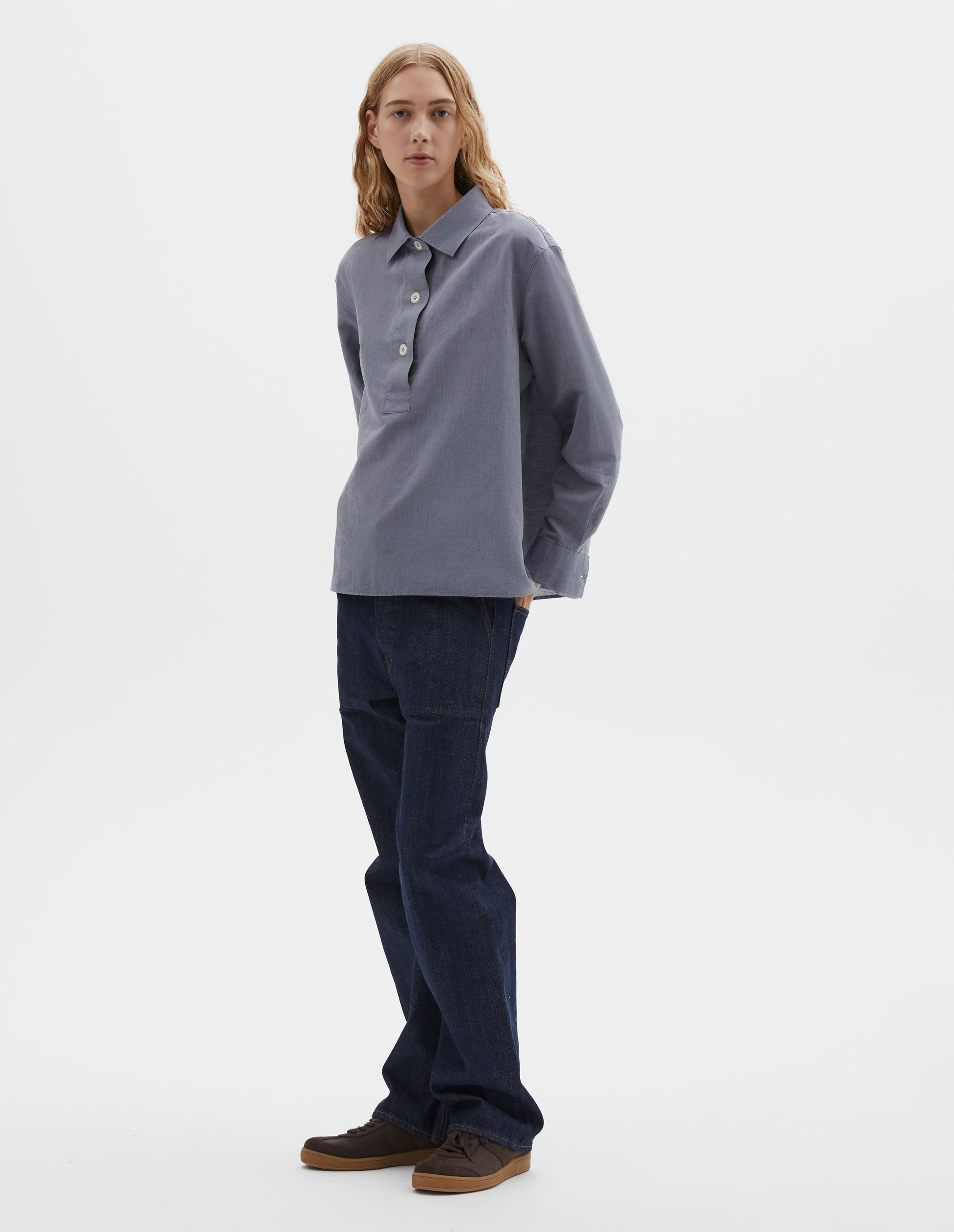 Full body image, front view of model wearing greyish blue long sleeve cotton linen shirt, with half raised placket and corozo brace buttons. Styled with indigo blue jeans and brown suede trainers.
