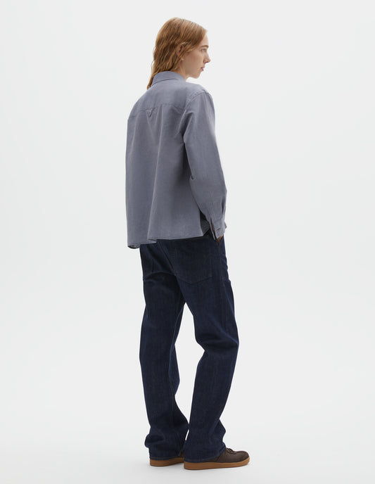 Full body image, back view of model wearing indigo blue denim twill jeans, with stitch-through waistband with belt loops, cinch back detail with metal buckle and patch back pockets. Styled with blue chambray shirt and brown suede trainers.
