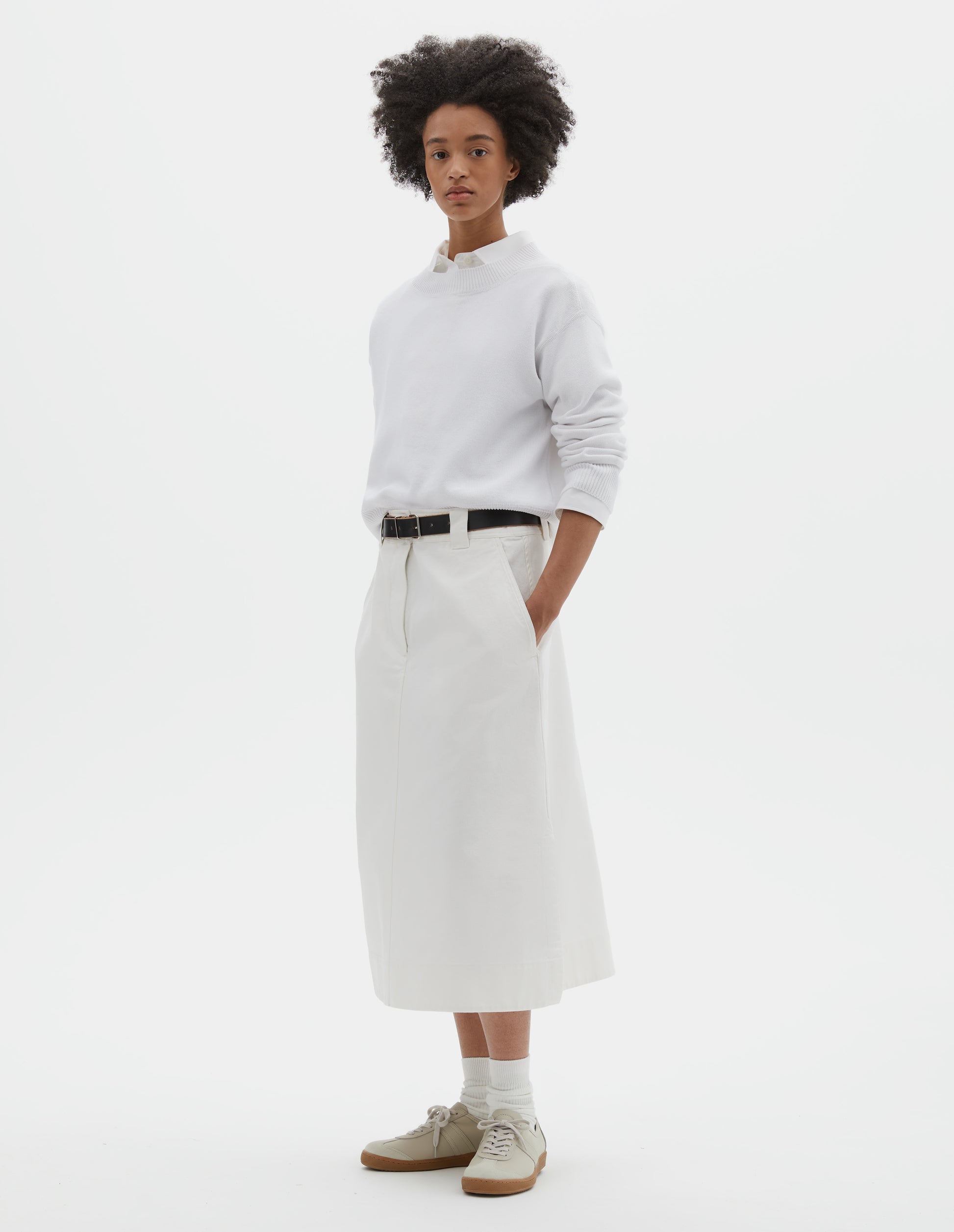 Full body image, front view of model wearing white regular cut long length A line skirt with side splits, zip fly and double hook and bar fastening waistband, belt loops, slant pockets and side splits. Styled with white jumper over white shirt, black leather belt, white socks and beige nubuck trainers