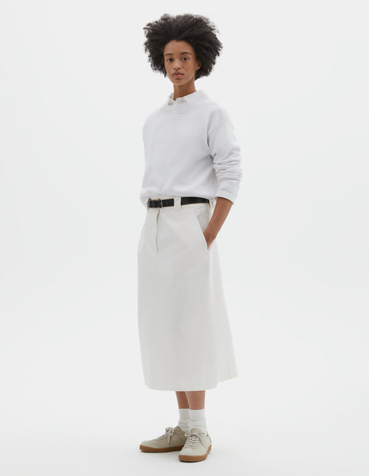 Full body image, front view of model wearing white regular cut long length A line skirt with side splits, zip fly and double hook and bar fastening waistband, belt loops, slant pockets and side splits. Styled with white jumper over white shirt, black leather belt, white socks and beige nubuck trainers