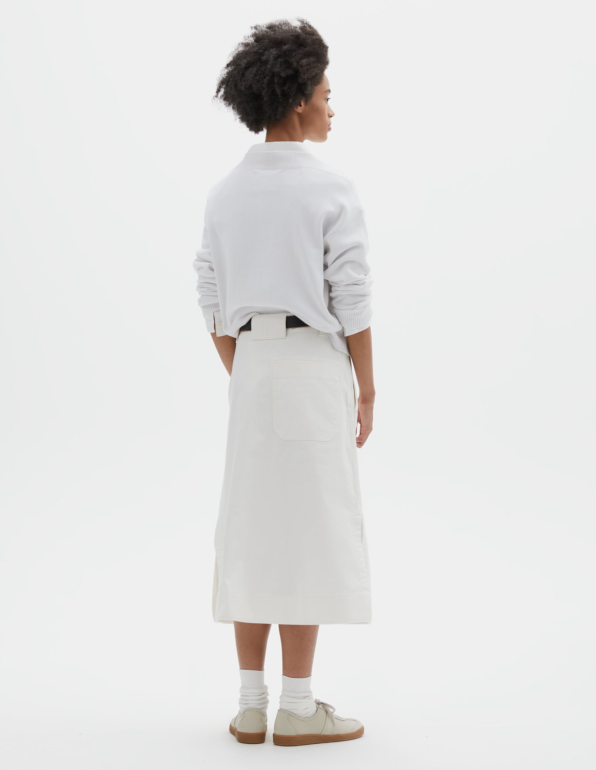 Full body image, back view of model wearing white regular cut long length A line skirt with side splits, belt loop detail on back and back patch pocket with reinforced stitch detailing. Styled with white jumper over white shirt, black leather belt, white socks and beige nubuck trainers. 