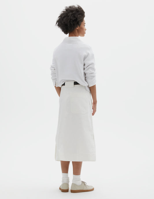 Full body image, back view of model wearing white regular cut long length A line skirt with side splits, belt loop detail on back and back patch pocket with reinforced stitch detailing. Styled with white jumper over white shirt, black leather belt, white socks and beige nubuck trainers. 