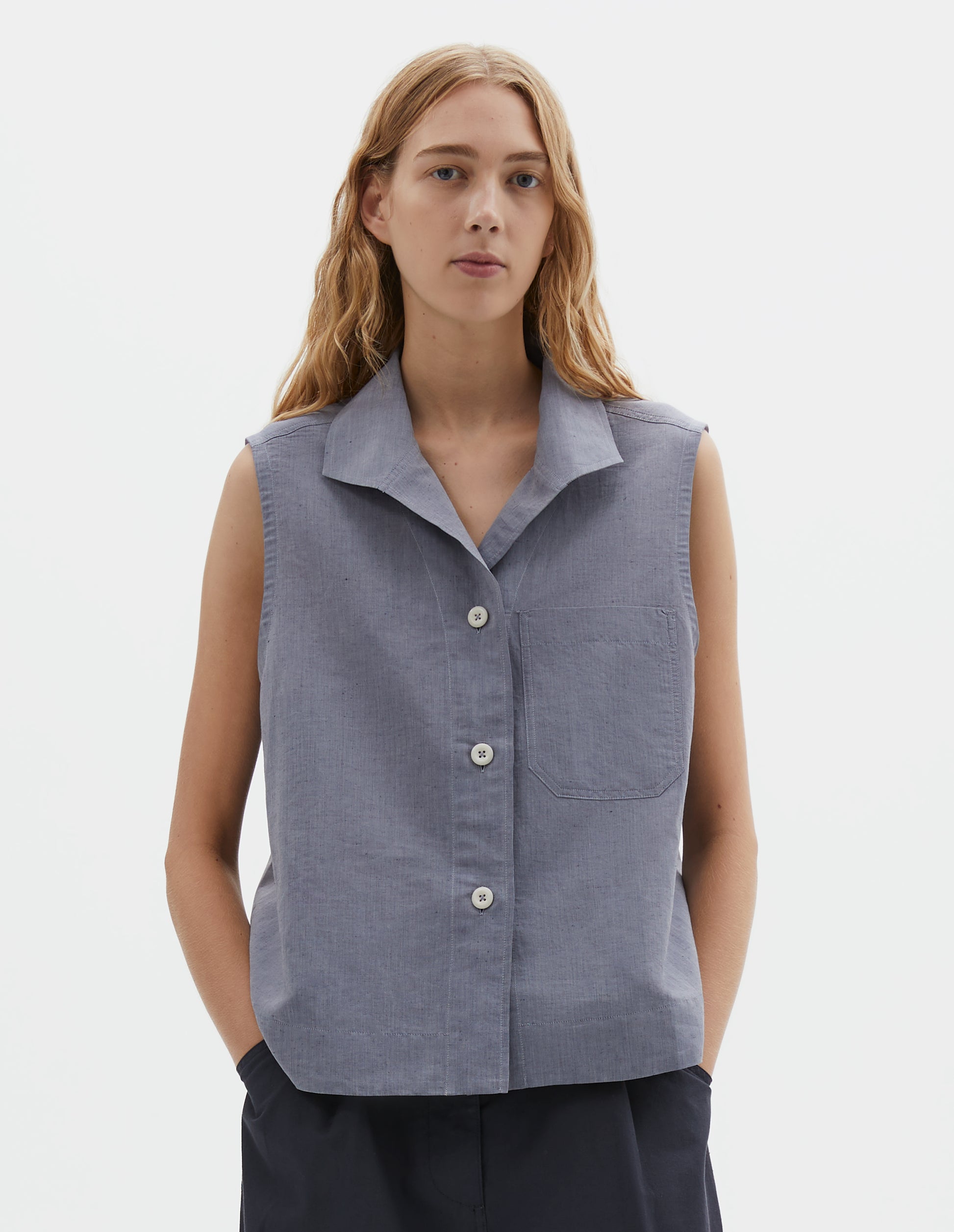 Cropped body image, front view of model wearing blueish grey cotton sleeveless shirt with a one-piece open spread collar, wide stitch-through placket, corozo brace button closure and single chest pocket. Styled with dark navy blue trousers. 