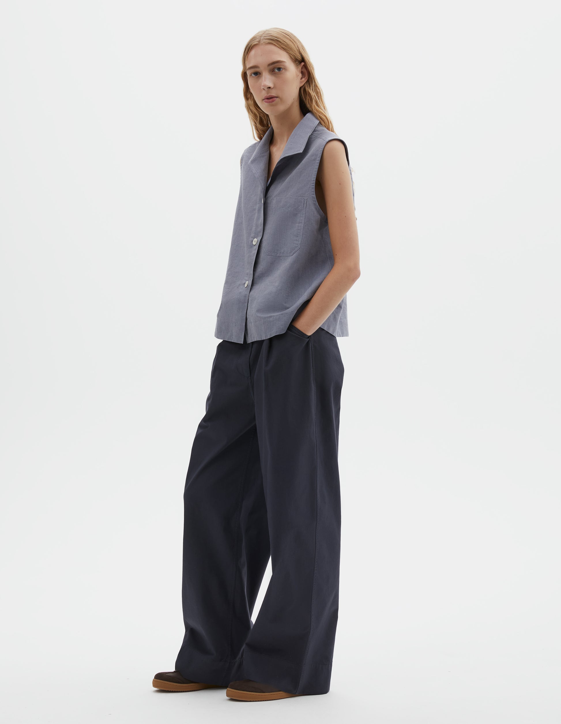Full body image, front view of model wearing blueish grey cotton sleeveless shirt with a one-piece open spread collar, wide stitch-through placket, corozo brace button closure and single chest pocket. Styled with dark navy blue trousers and brown suede trainers with gum sole. 
