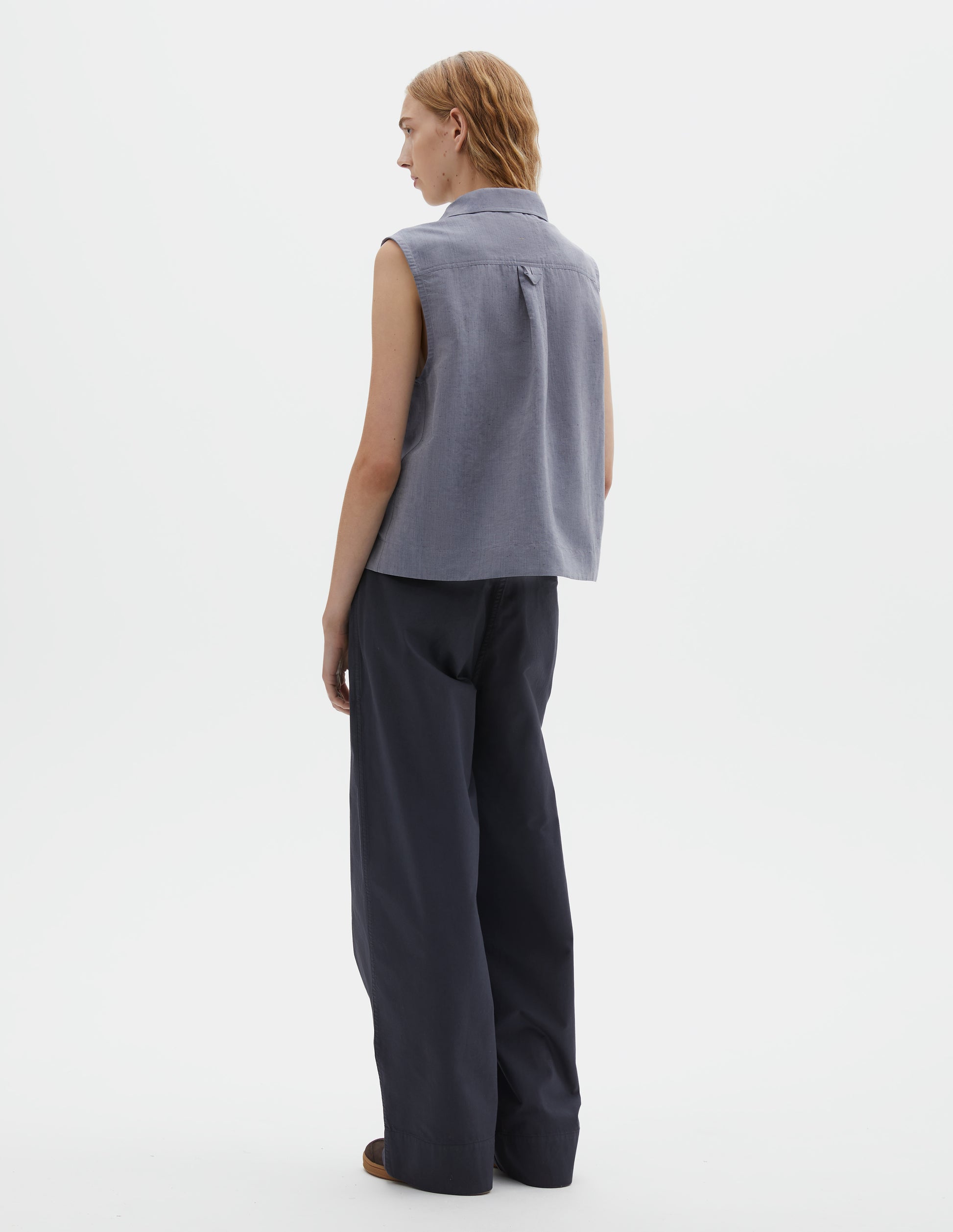 Full body image, back view of model wearing blueish grey cotton sleeveless shirt with a box pleat and hanger loop detail on back yoke. Styled with dark navy blue trousers and brown suede trainers with gum sole.