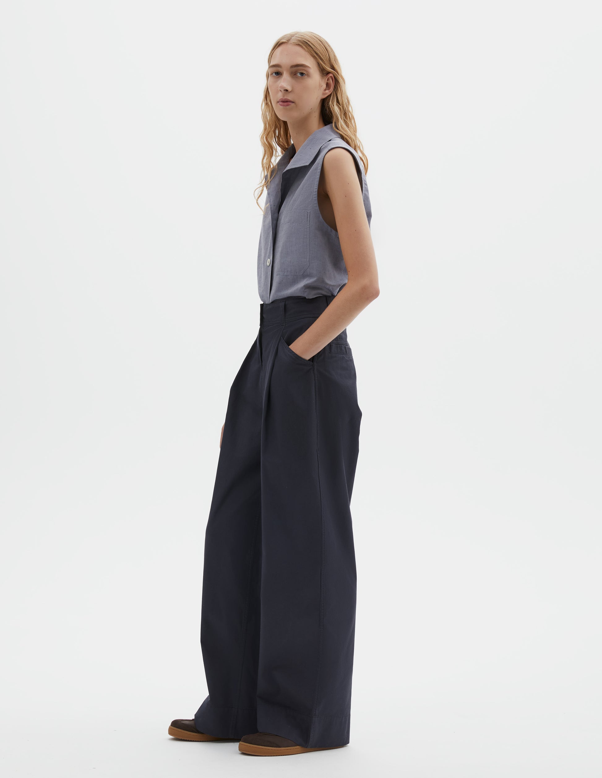 Full body image, front view of model wearing dark navy wide leg pleated trousers, with high waist, deep pleats and slant pockets. Styled with light blue sleeveless shirt and brown suede trainers.
