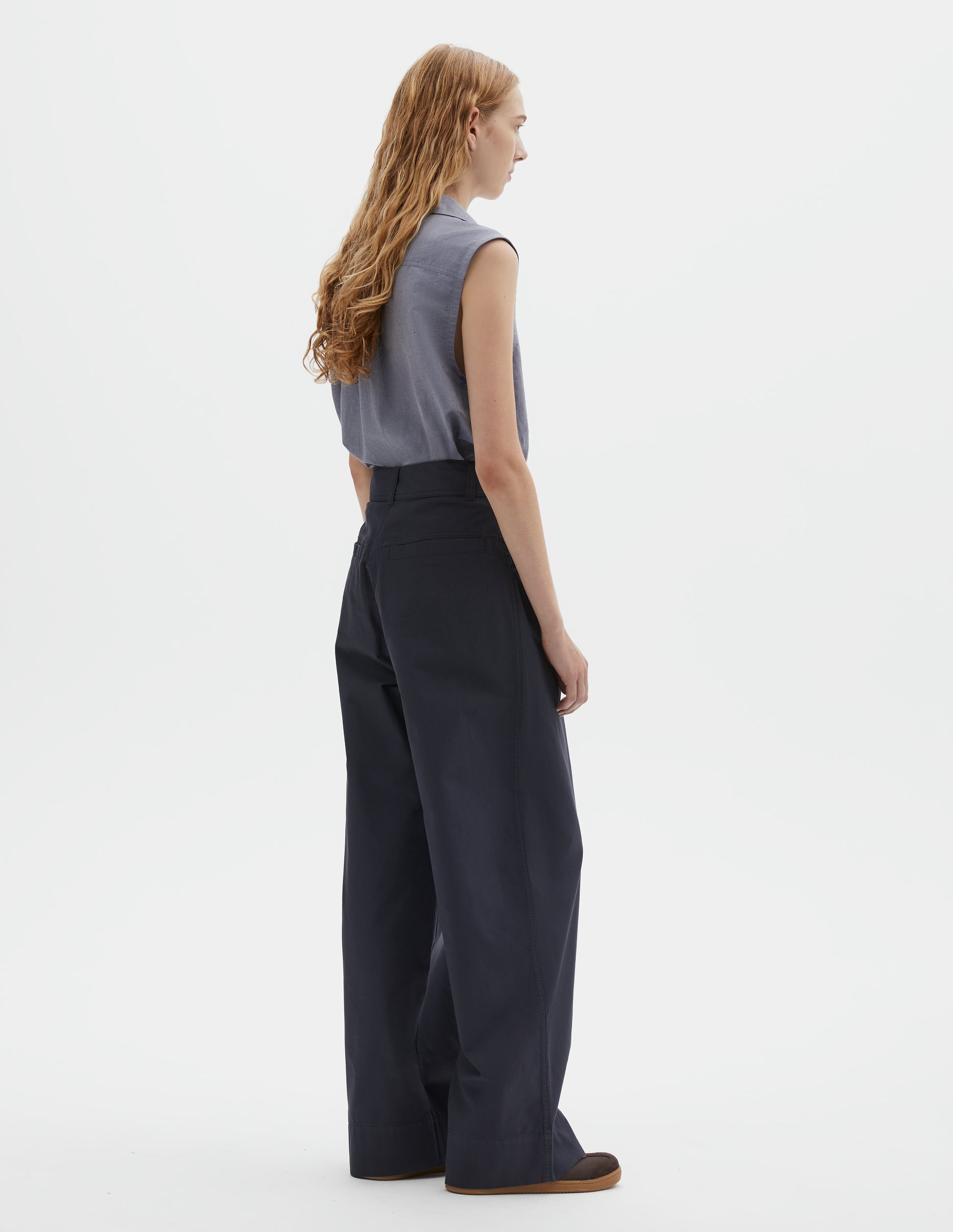 Full body image, back view of model wearing dark navy wide leg pleated trousers, with high waist, belt loops and welt back pockets. Styled with light blue sleeveless shirt and brown suede trainers.