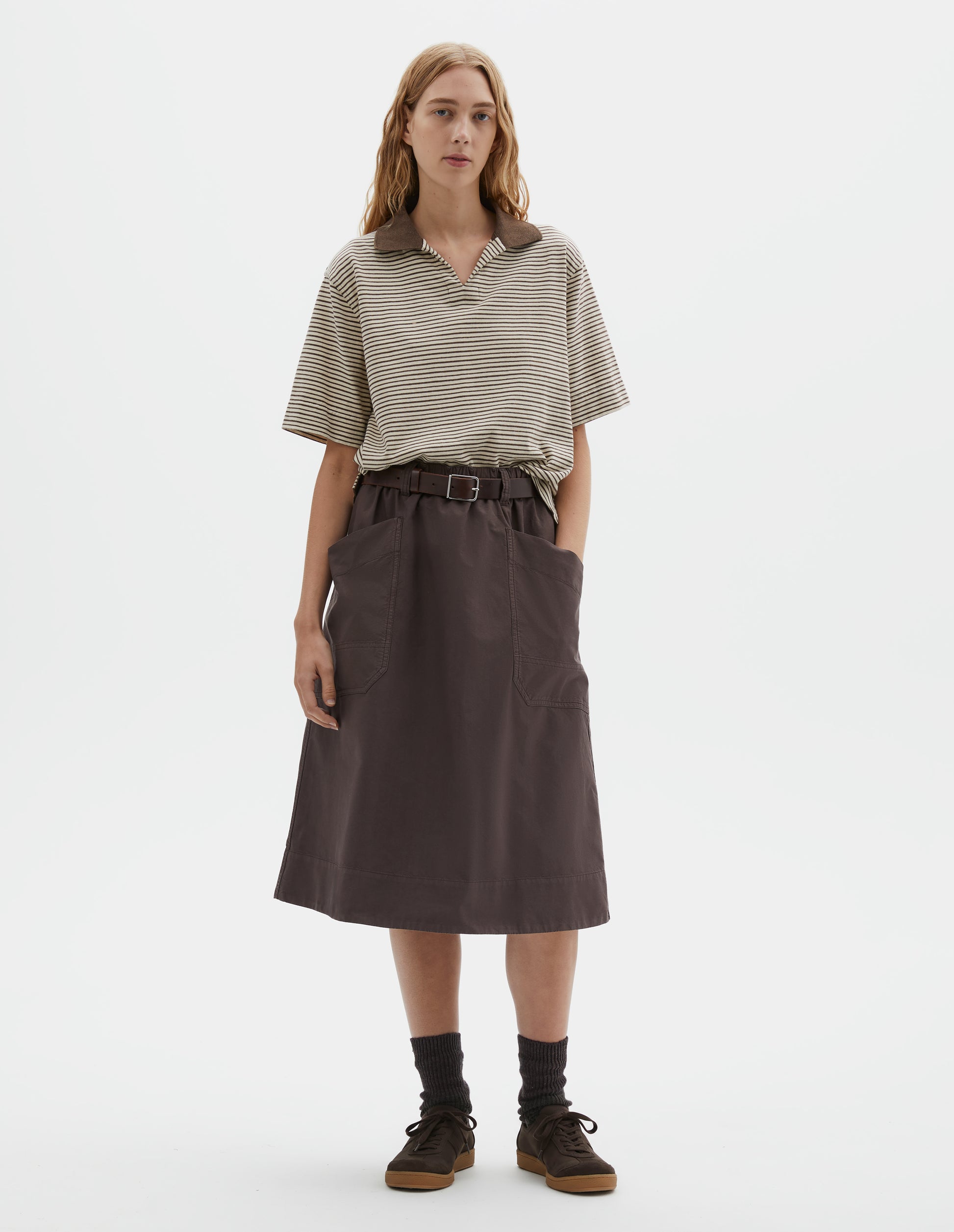 Full body image, front view of model wearing brown cotton skirt with large slant patch pockets, elasticated waistband with belt loops and deep stitch hem. Styled with brown and white striped short sleeve polo shirt, brown leather belt, black socks and brown suede trainers.