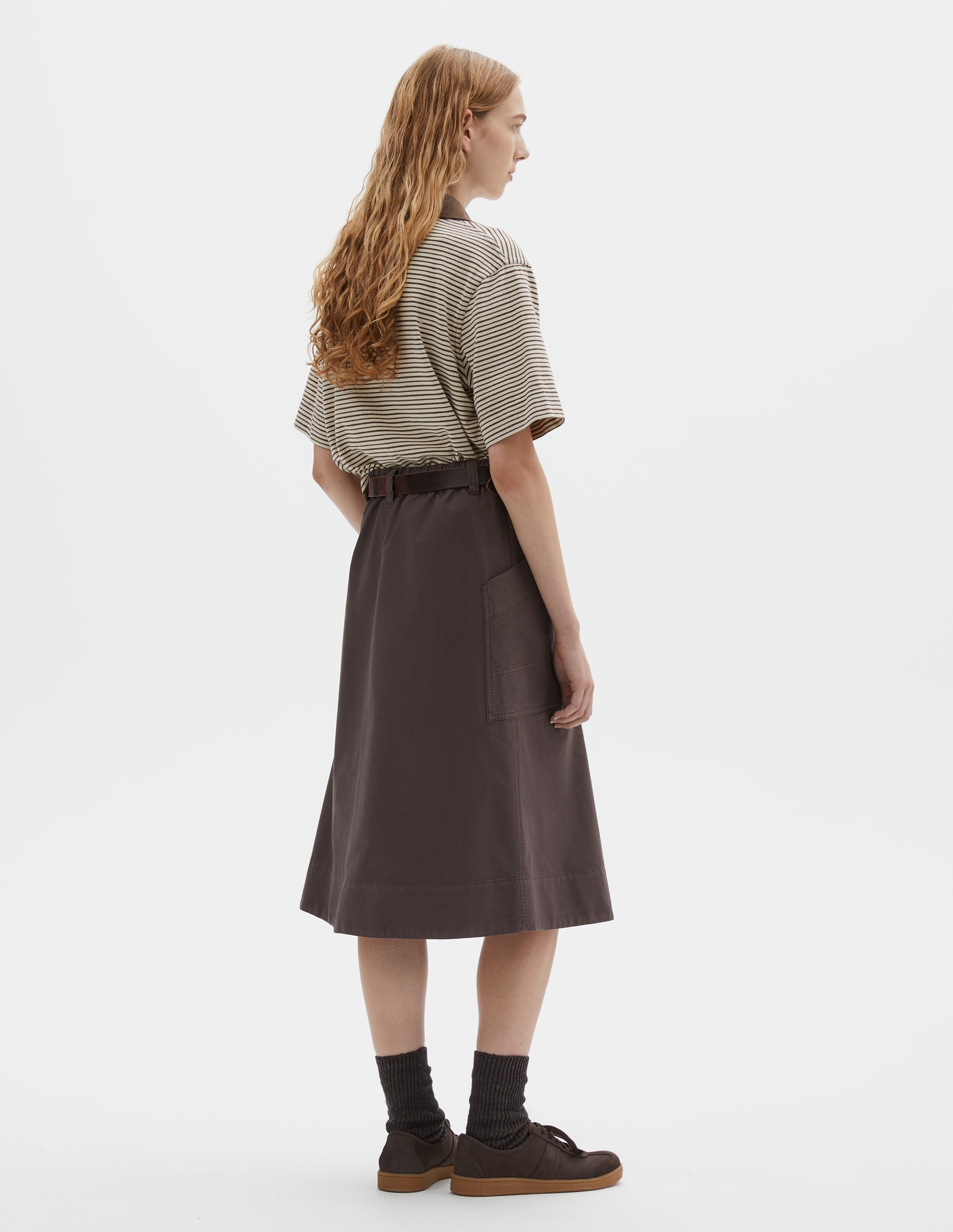 Full body image, back view of model wearing brown cotton skirt with large slant patch pockets, elasticated waistband with belt loops and deep stitch hem. Styled with brown and white striped short sleeve polo shirt, brown leather belt, black socks and brown suede trainers. 