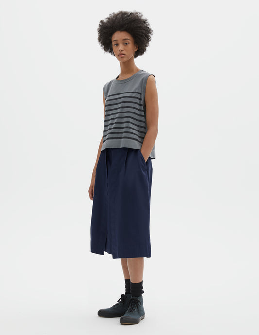Full body image, front view of model wearing blue cotton linen tank top with black stripes, contrast ribbing on neckline and armholes, with side splits and deep stitch hem. Styled with blue skirt, black socks and blue canvas high top trainers.
