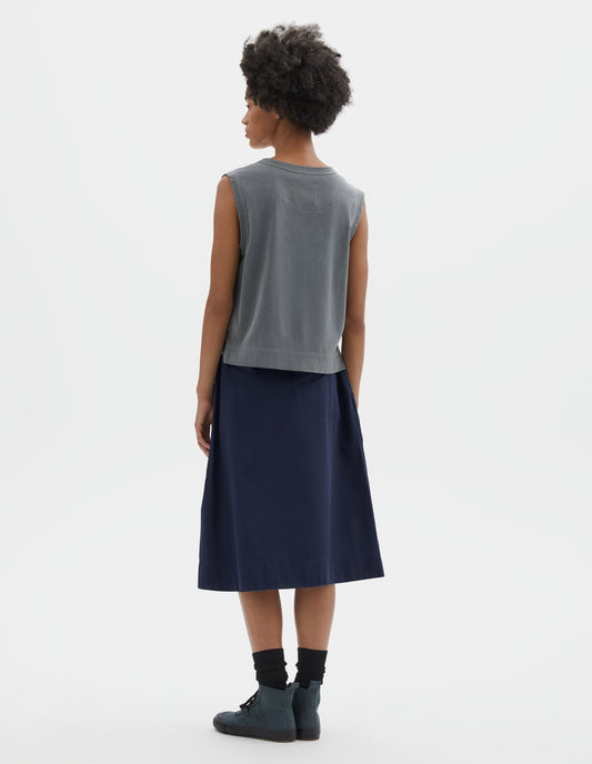 Full body image, back view of model wearing blue cotton linen tank top with contrast ribbing on neckline and armholes, with stitch-through half moon on back yoke, side splits and deep stitch hem. Styled with blue skirt, black socks and blue canvas high top trainers.