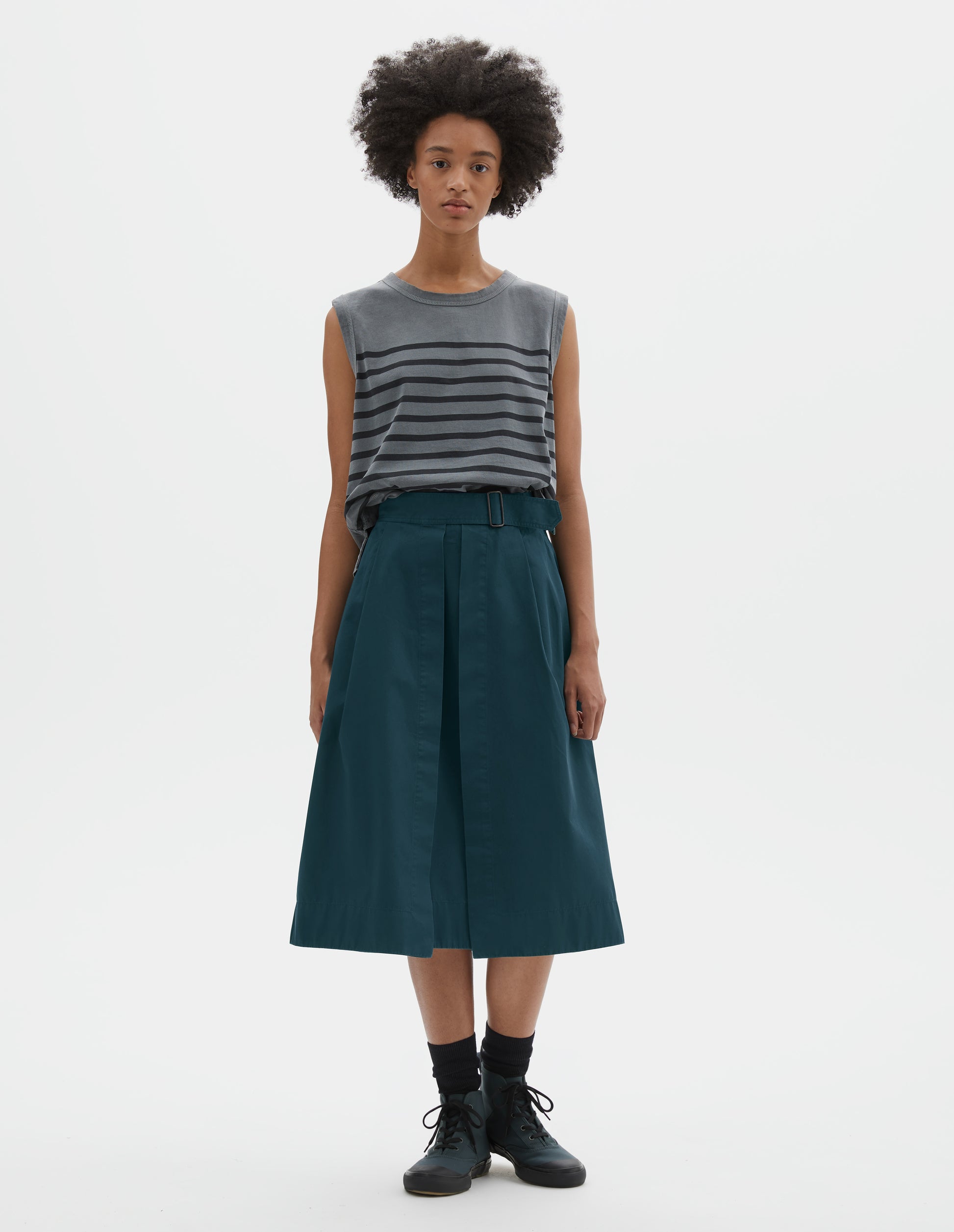 Full body image, front view of model wearing greenish blue cotton twill skirt, with wide crossover waistband, button fastening slide adjuster and slant pockets. Styled with sleeveless blue and black striped vest, black socks and blue canvas trainers.
