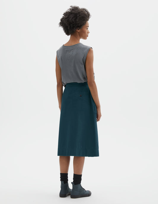 Full body image, back view of model wearing greenish blue cotton twill skirt, with wide crossover waistband and stitch through welt back pocket. Styled with sleeveless blue and black striped vest, black socks and blue canvas trainers. 