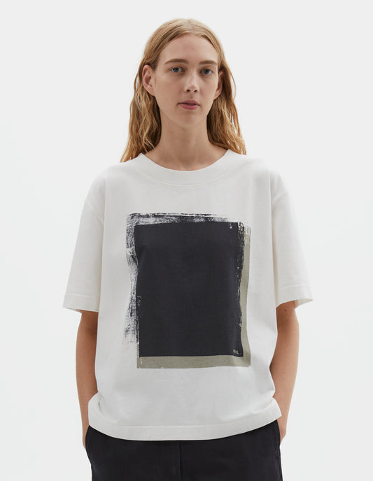 Cropped body image, front view of model wearing off white short sleeve t shirt with black abstract block printed on the front. Styled with black tapered trousers.