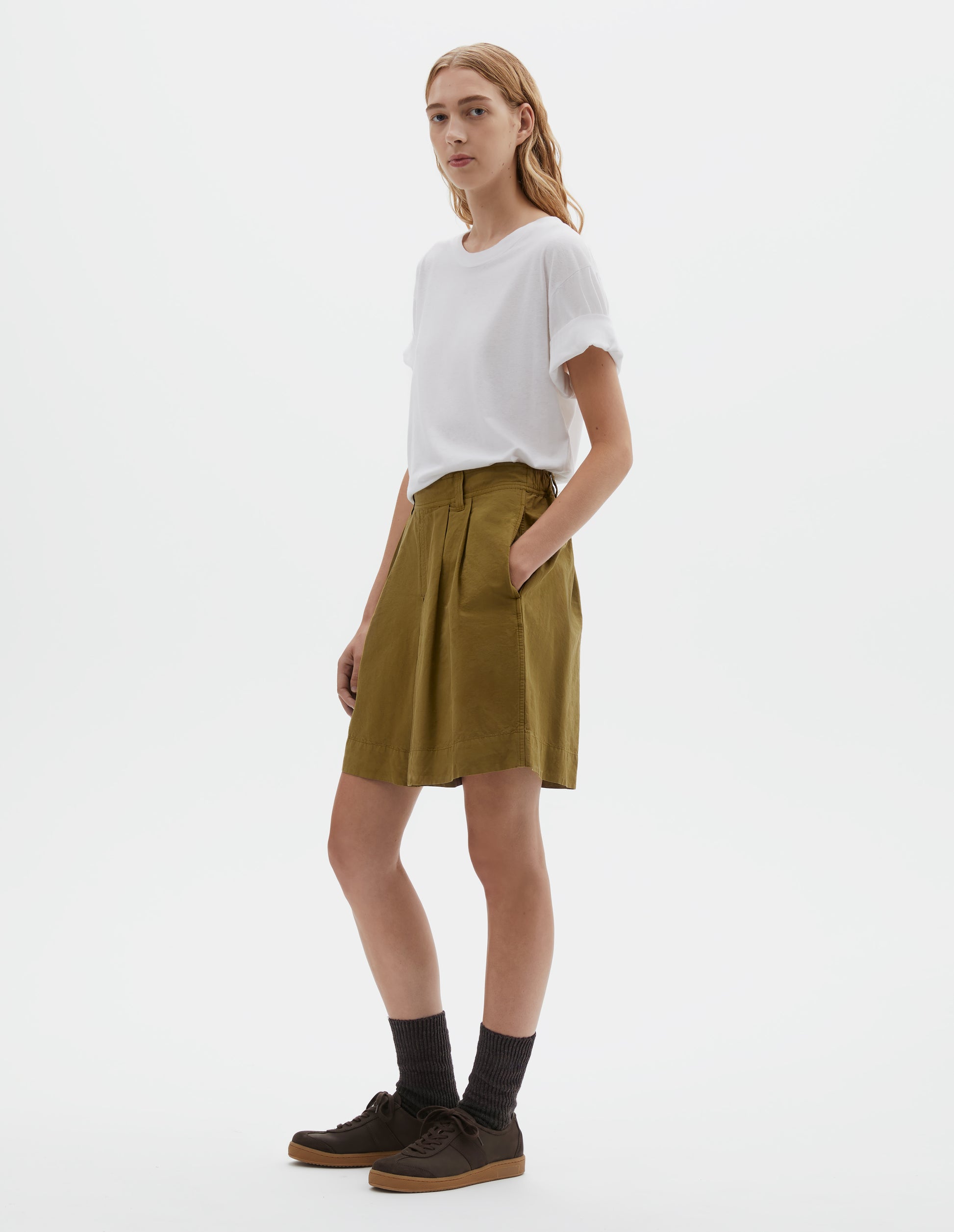 Full body image, front view of model wearing yellowish green cotton linen shorts with pleated skirt front panel, semi-elasticated waistband, side pockets, and belt loops. Styled with white t-shirt, dark brown socks and dark brown nubuck trainers.