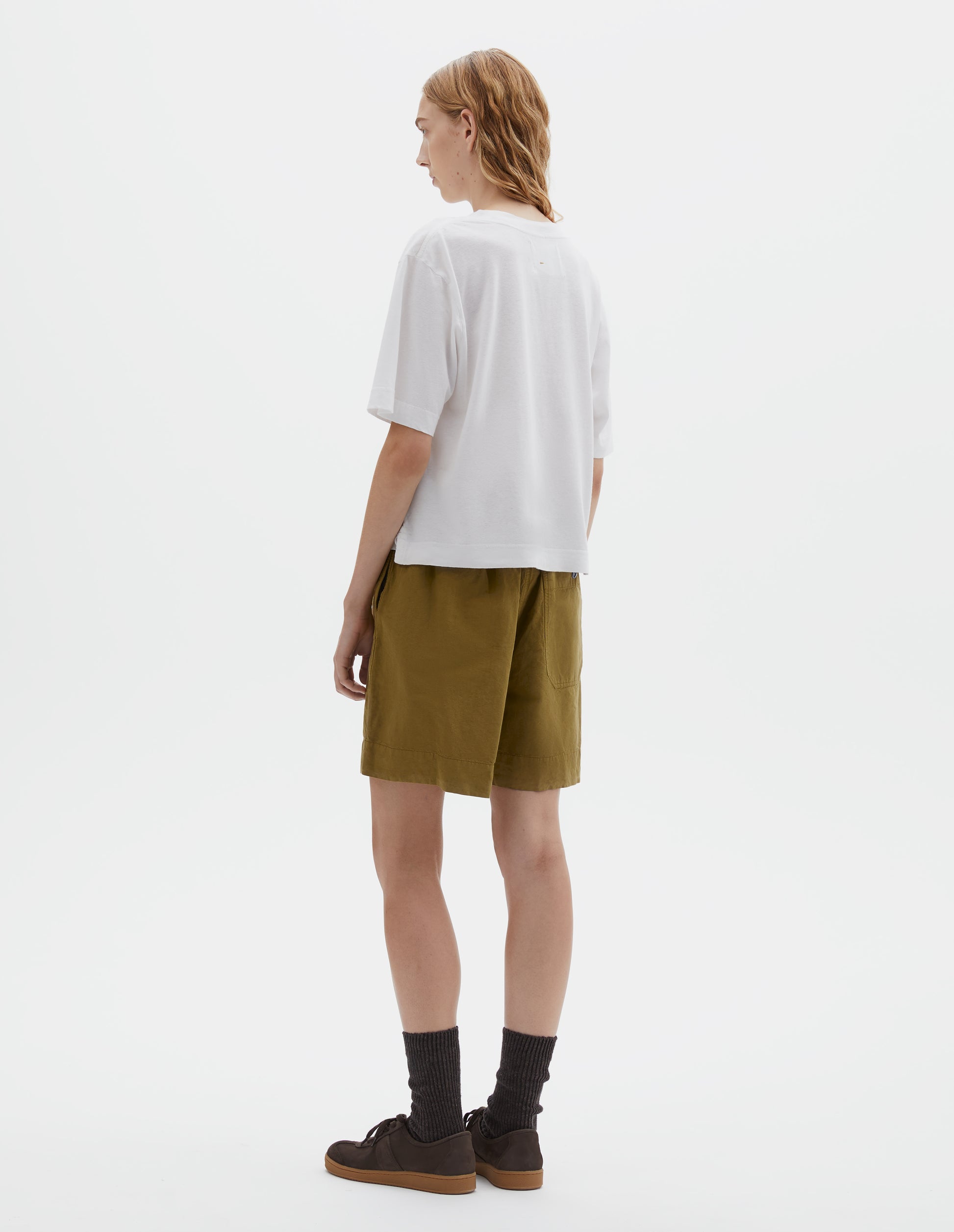 Full body image, back view of model wearing white short sleeve cotton t-shirt with contrast rib on neck and stitch detailing on back shoulder. Styled with olive green shorts, grey socks and brown suede trainers.