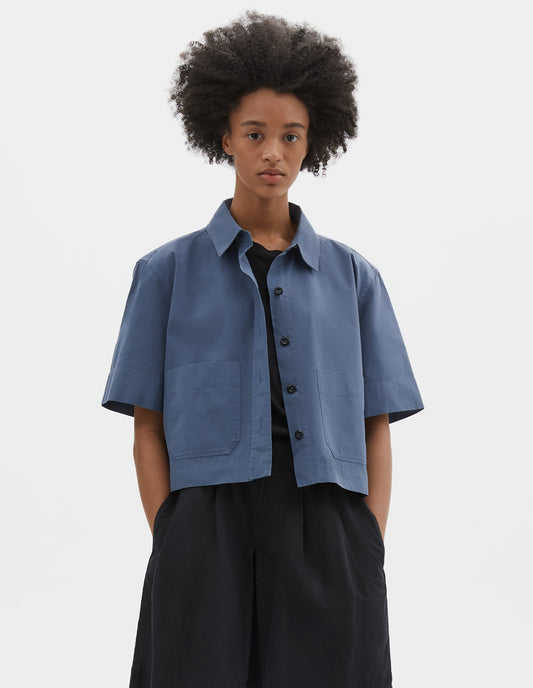 Cropped body image, front view of model wearing blue cotton short sleeve shirt, with raised placket, patch pockets, stepped side hem and corozo brace buttons. Styled over black t-shirt and black pleated shorts.