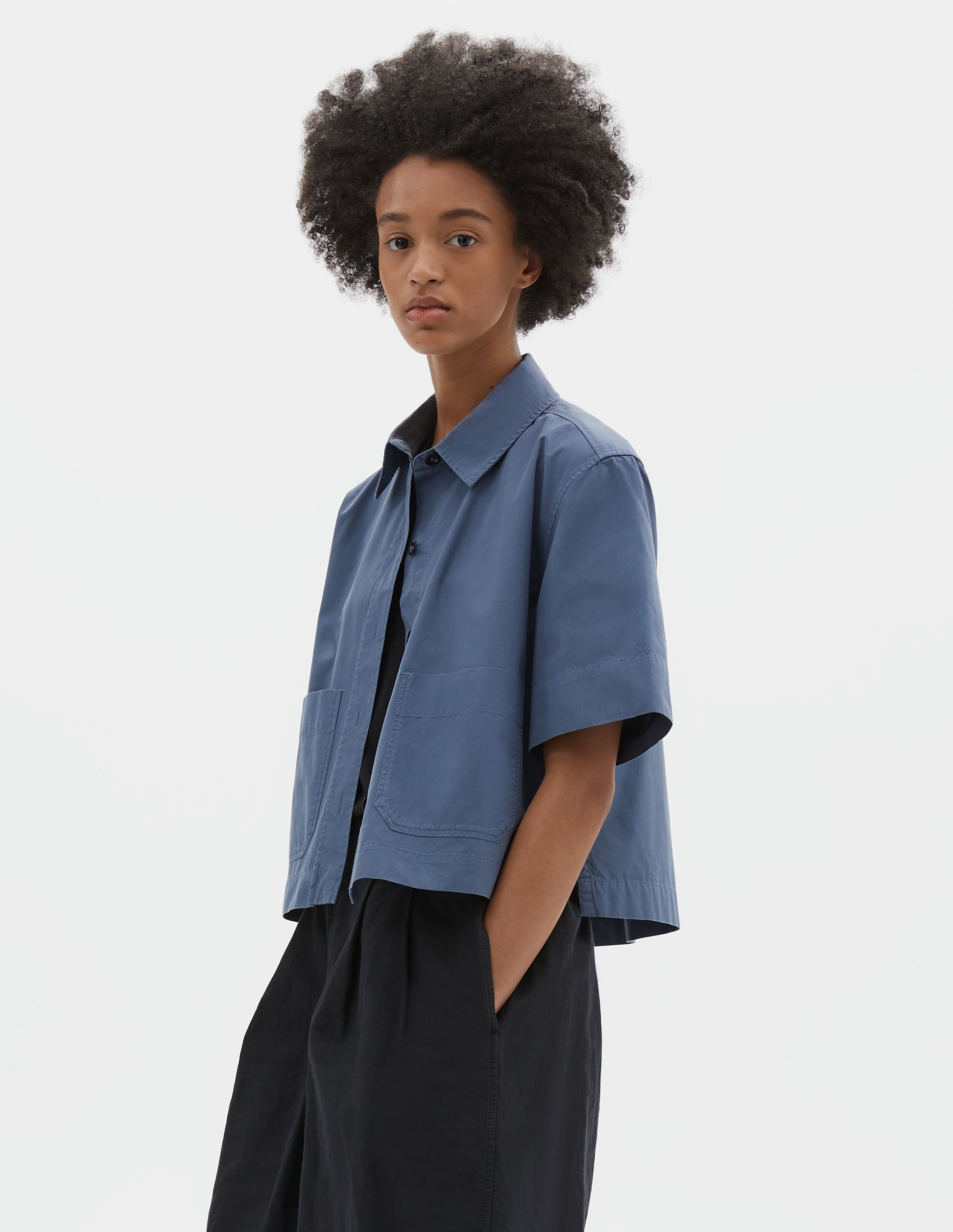 Cropped body image, side view of model wearing blue cotton short sleeve shirt, with raised placket, patch pockets, stepped side hem and corozo brace buttons. Styled over black t-shirt and black pleated shorts.