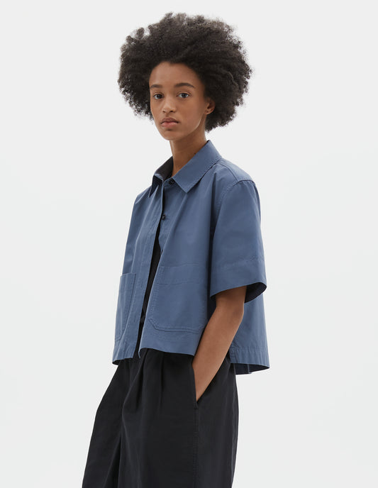 Cropped body image, side view of model wearing blue cotton short sleeve shirt, with raised placket, patch pockets, stepped side hem and corozo brace buttons. Styled over black t-shirt and black pleated shorts.