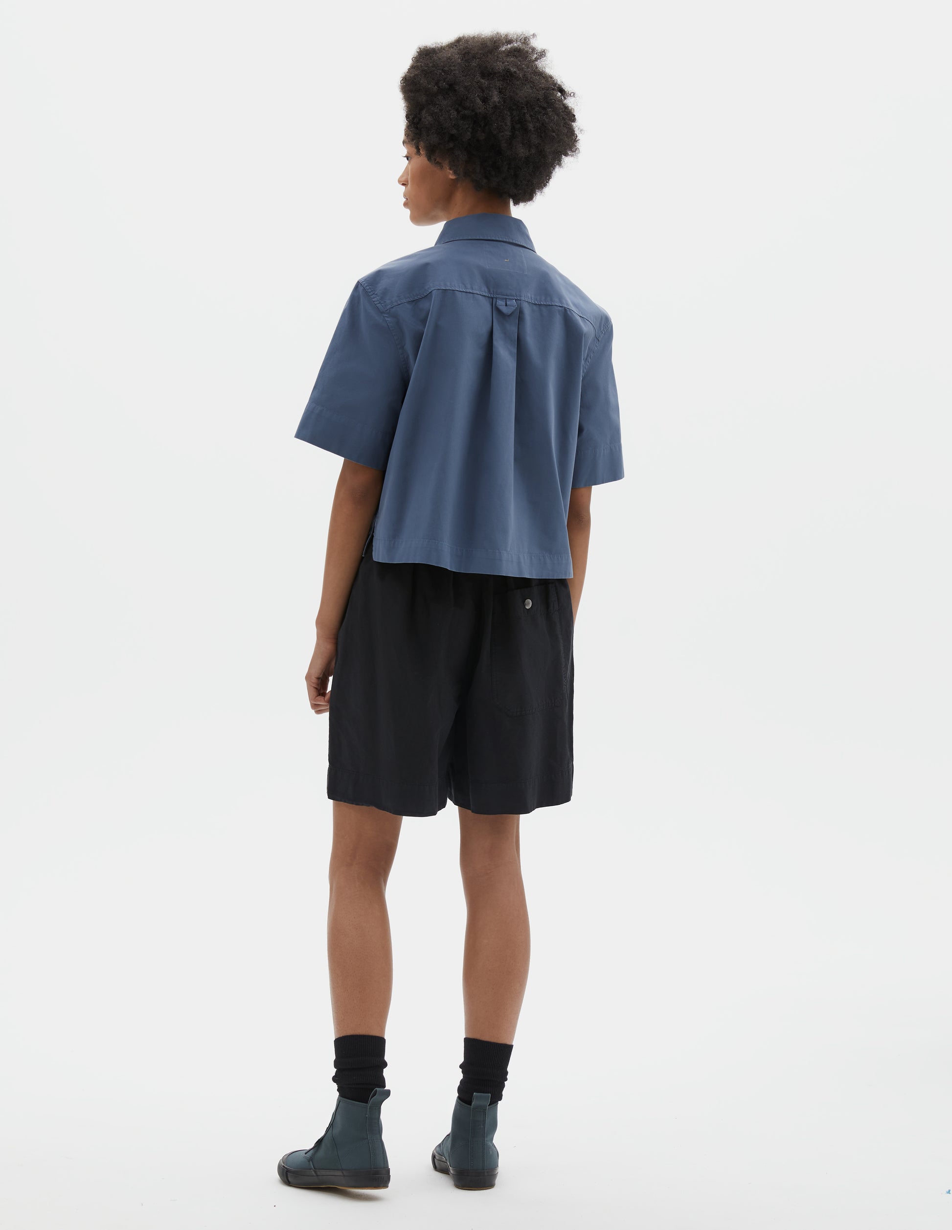 Full body image, back view of model wearing blue cotton short sleeve shirt, with stepped side hem, hanger loop and box pleat on back yoke. Styled with black shorts, black socks and blue canvas high top trainers.