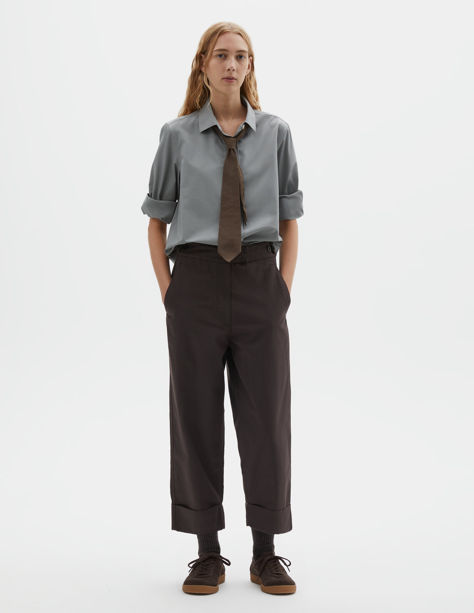 Full body image, front view of model wearing dark brown cotton drill trousers with slant pockets, corozo button detailing on waistband and turn up hems. Styled with grey shirt with rolled up sleeves, brown tie and dark brown socks with dark brown nubuck trainers.