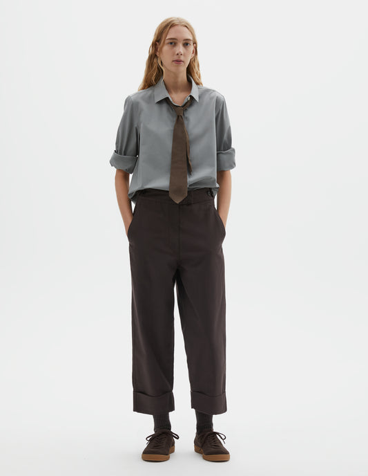 Full body image, front view of model wearing dark brown cotton drill trousers with slant pockets, corozo button detailing on waistband and turn up hems. Styled with grey shirt with rolled up sleeves, brown tie and dark brown socks with dark brown nubuck trainers.