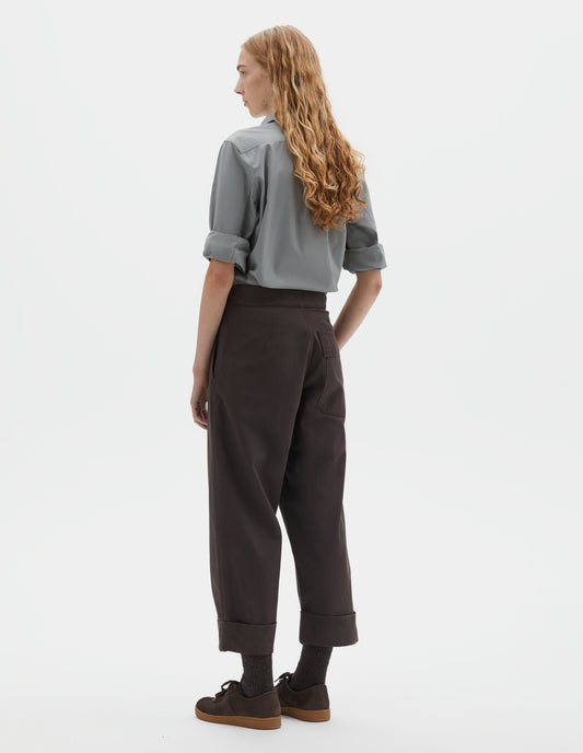 Full body image, back view of model wearing dark brown cotton drill trousers with adjustable tab detailing on waistband and flap patch pocket. Styled with grey shirt with rolled up sleeves and dark brown socks with dark brown nubuck trainers.