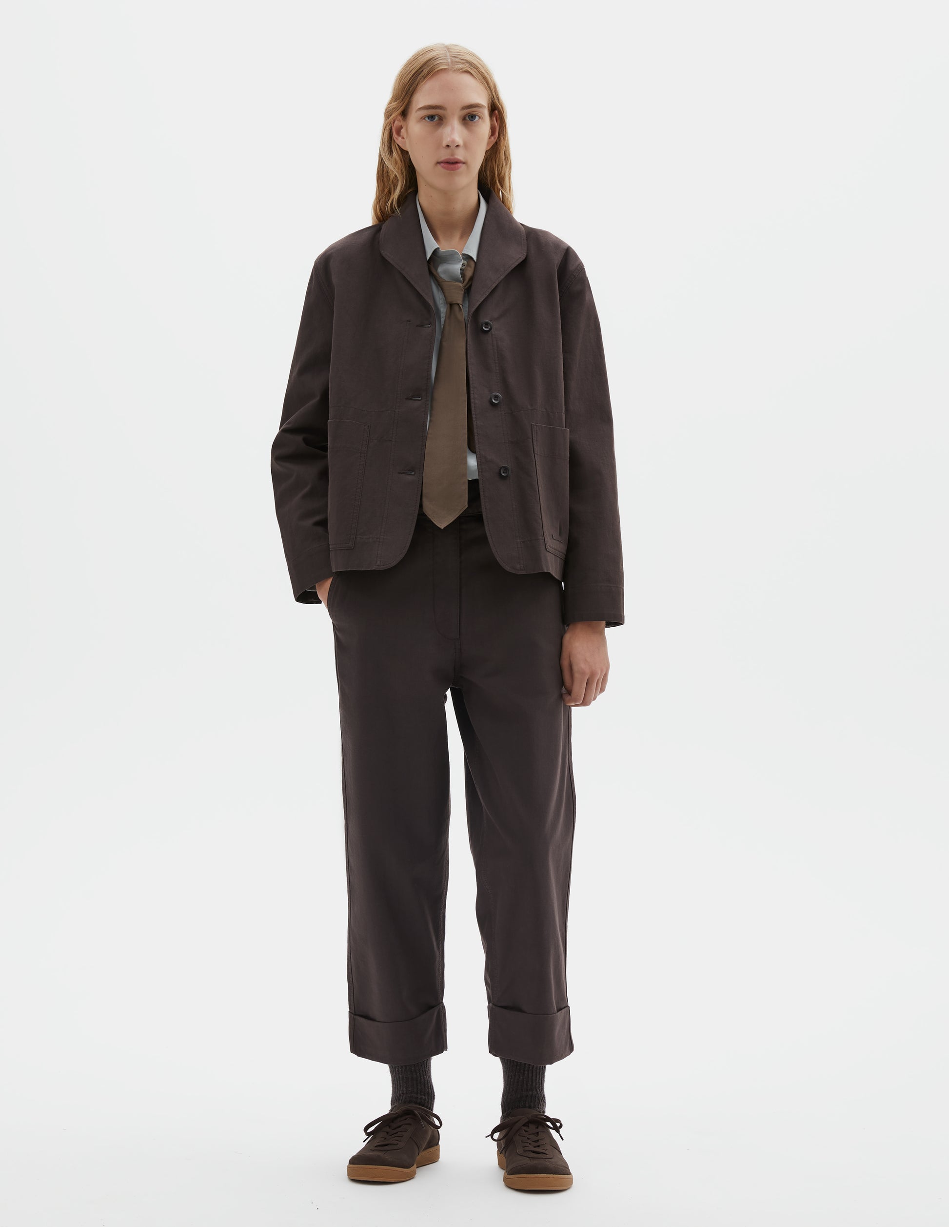 Full body image, front view of model wearing dark brown cotton drill jacket with two patch pockets, corozo button closure and collar. Styled with grey shirt, brown tie and matching brown trousers, dark brown socks with dark brown nubuck trainers.
