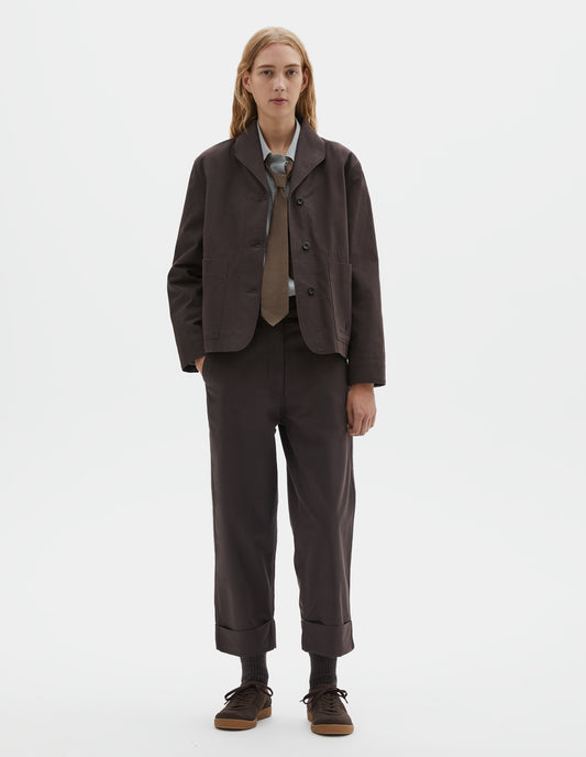 Full body image, front view of model wearing dark brown cotton drill jacket with two patch pockets, corozo button closure and collar. Styled with grey shirt, brown tie and matching brown trousers, dark brown socks with dark brown nubuck trainers.