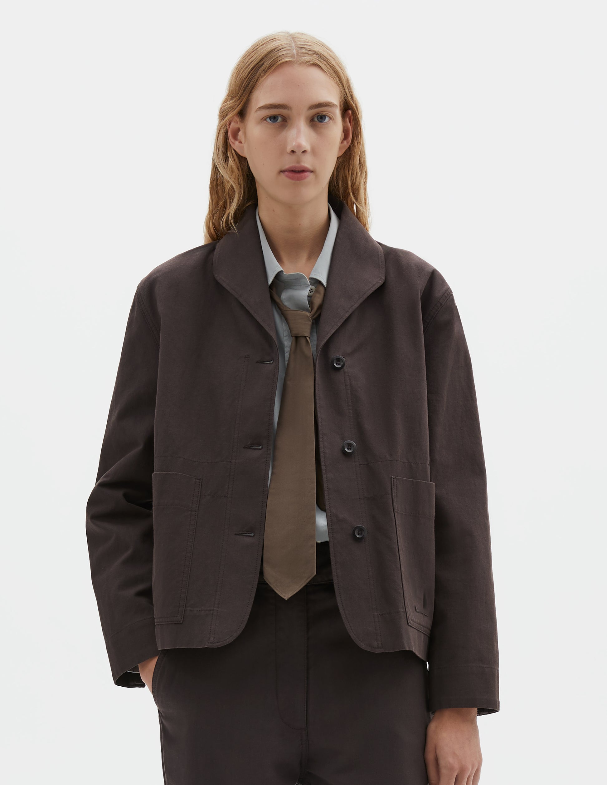 Cropped body image, front view of model wearing dark brown cotton drill jacket with two patch pockets, corozo button closure and collar. Styled with grey shirt, brown tie and matching brown trousers.
