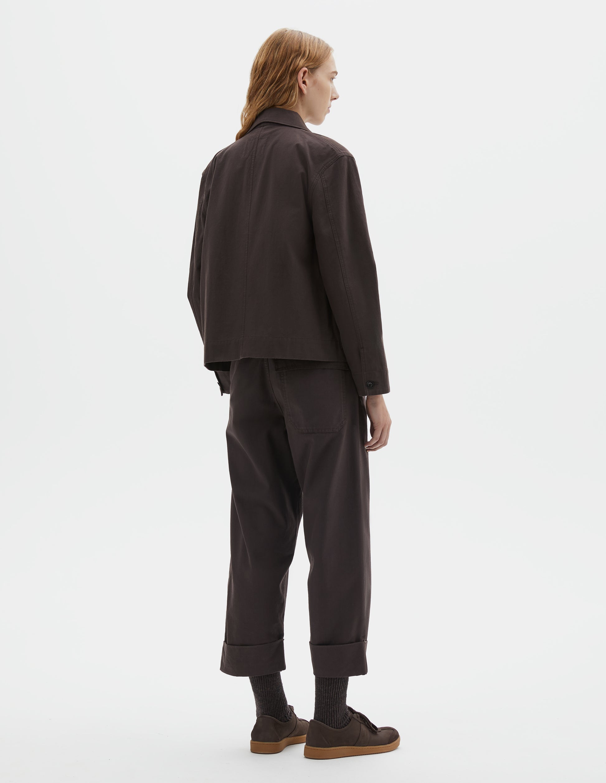 Full body image, back view of model wearing dark brown cotton drill jacket with corozo button cuffs. Styled with matching brown trousers, dark brown socks and dark brown nubuck trainers.