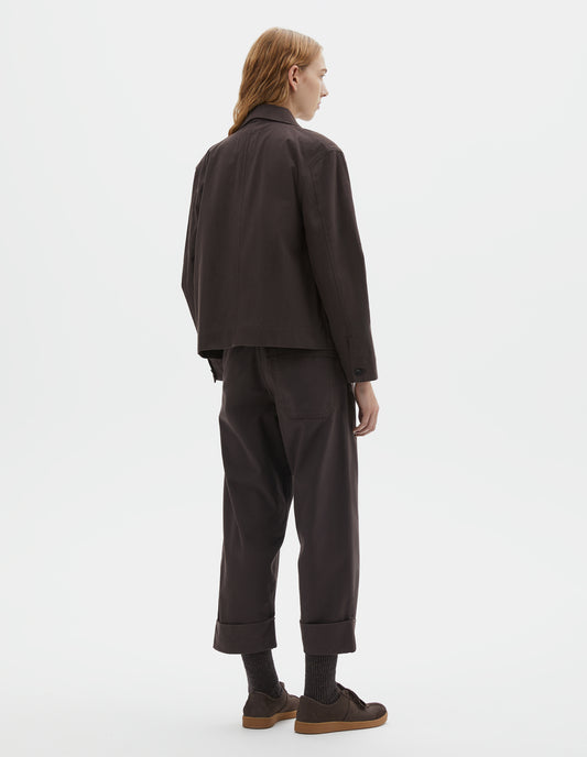 Full body image, back view of model wearing dark brown cotton drill jacket with corozo button cuffs. Styled with matching brown trousers, dark brown socks and dark brown nubuck trainers.