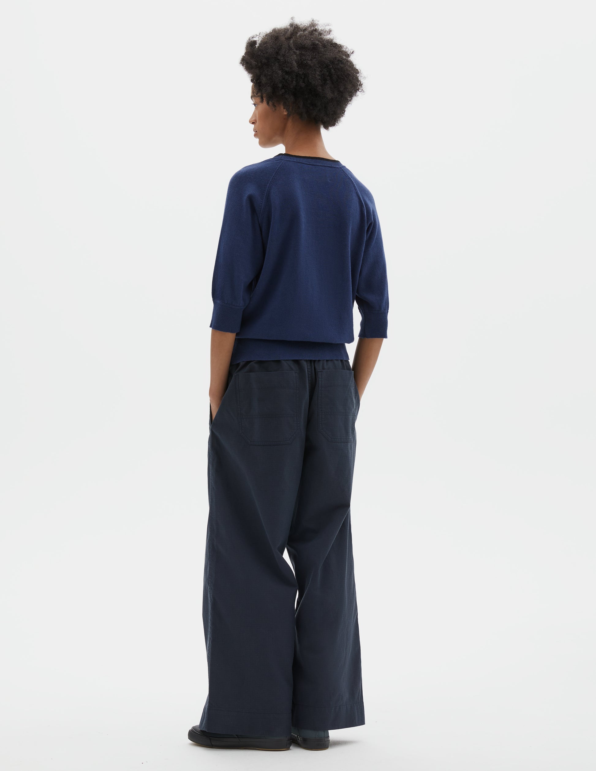 Full body image, back view of model wearing blue short sleeve knitted t-shirt, with contrast black stitching on neckline, raglan sleeve, flatlock stitch seam detailing and contrast rib on sleeve and hem. Styled with navy blue wide leg joggers and blue canvas trainers.