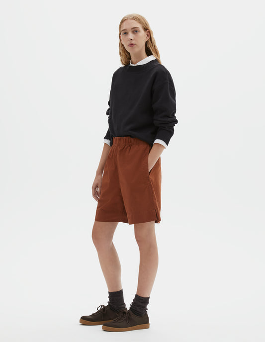 Full body image, front view of model wearing dark orange relaxed cut shorts with elasticated waist, side pockets and side split hems. Styled with black jumper over white shirt and dark brown socks with dark brown nubuck trainers.