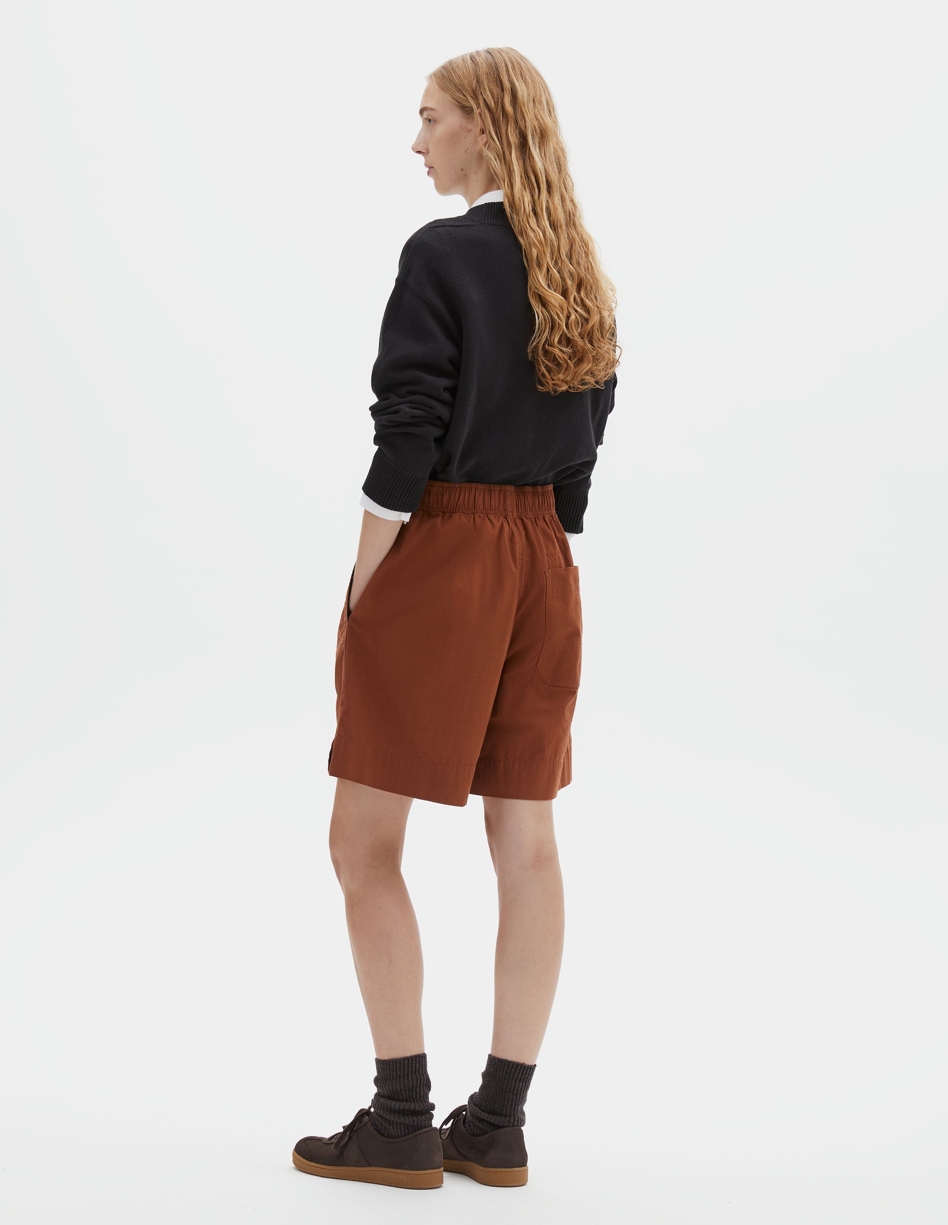 Full body image, back view of model wearing dark orange relaxed shorts with elasticated waist, side pockets and button-through patch pocket. Styled with black jumper over white shirt and dark brown socks with dark brown nubuck trainers.