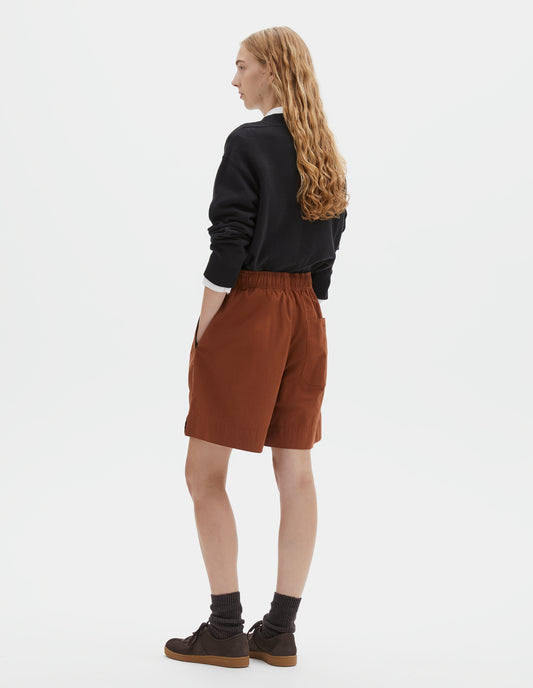 Full body image, back view of model wearing dark orange relaxed shorts with elasticated waist, side pockets and button-through patch pocket. Styled with black jumper over white shirt and dark brown socks with dark brown nubuck trainers.