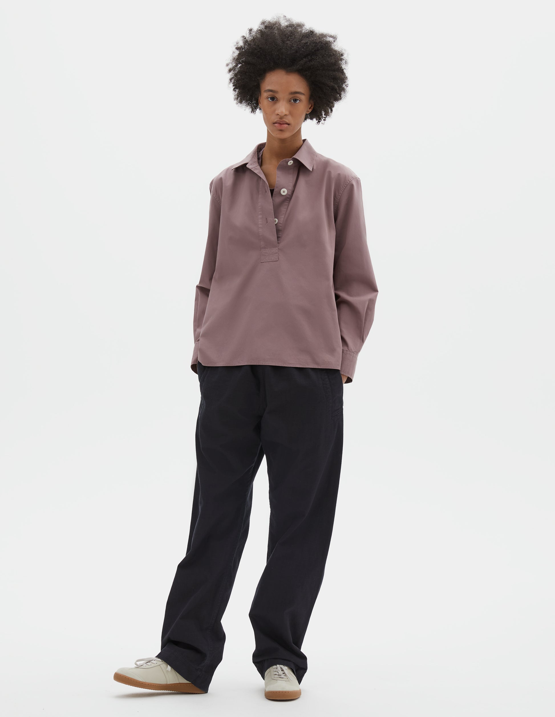 Full body image, front view of model wearing purpleish pink cotton poplin swing shirt, with half raised placket and corozo button fastening. Styled with dark navy trousers and beige suede trainers. 